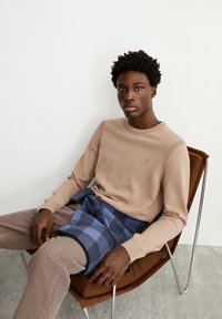 Beige Strickpullover mit Rundhalsausschnitt, kombiniert mit karierten Shorts in Blau und Schwarz. Model sitzt in einem braunen Stuhl auf einem grauen Boden.