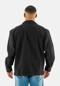 Chaqueta de tela negra y pesada, con mangas largas, un cuello y un corte recto. Presenta detalles estilo camisa en la parte posterior. Jeans azules visibles.