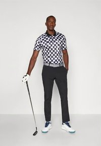 Koszulka polo w czarno-białą kratę z kołnierzykiem, w połączeniu z czarnymi spodniami. Białe buty do golfa z niebieskimi akcentami i kij golfowy w ręku.