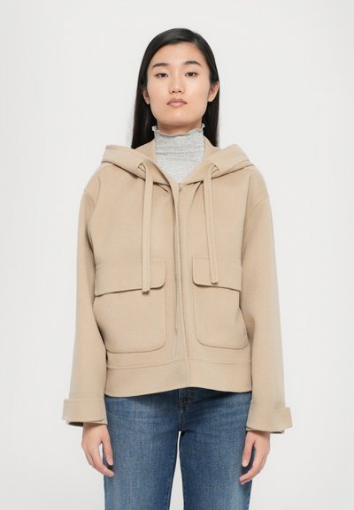 Veste à capuche beige en tissu doux avec deux poches avant, sécurisées par des rabats. Présente une coupe droite et des poignets retroussés.
