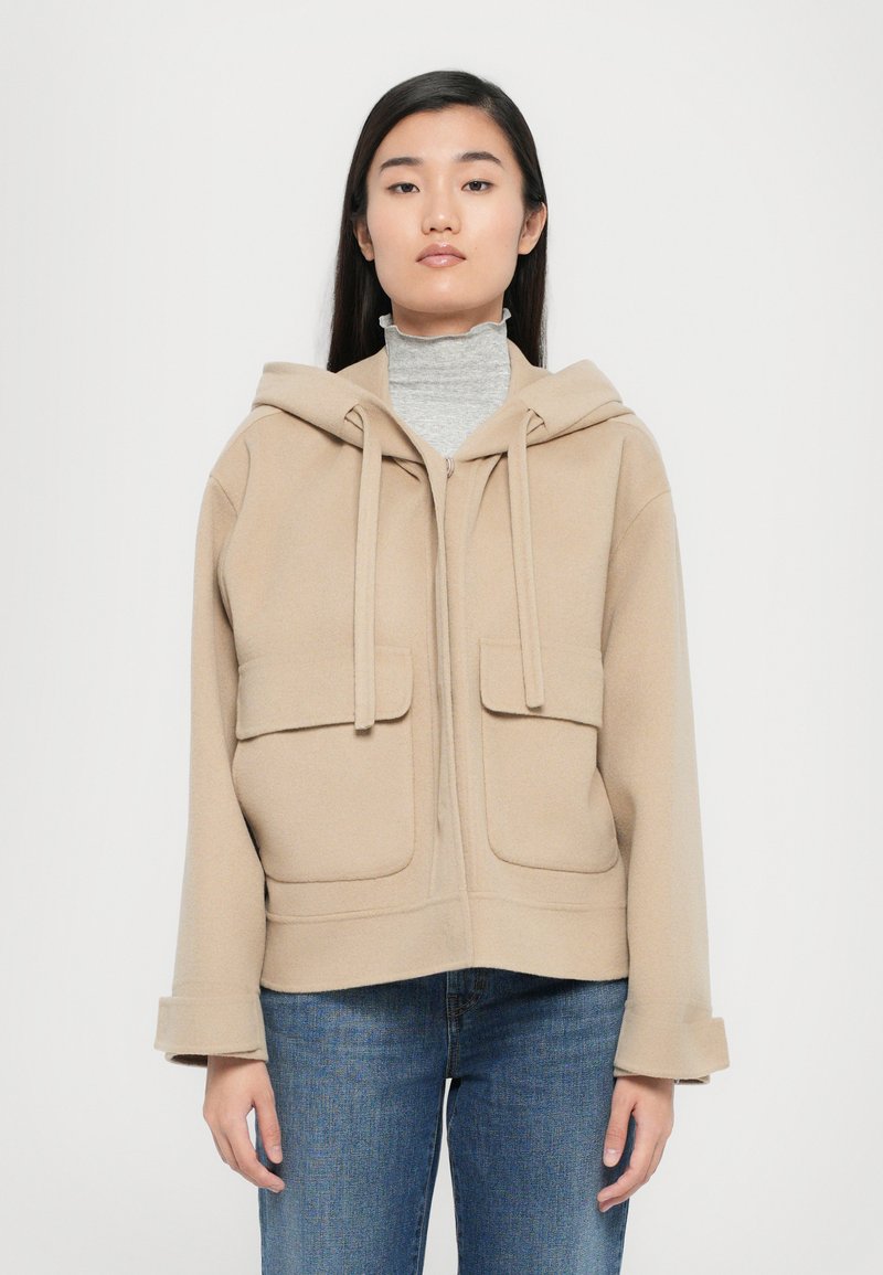 Veste à capuche beige en tissu doux avec deux poches avant, sécurisées par des rabats. Présente une coupe droite et des poignets retroussés.