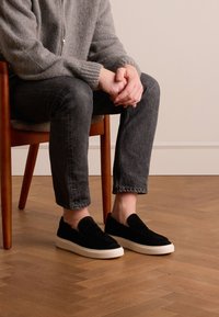 Black suede slip-on shoes with a white rubber sole, paired with gray denim jeans and a gray sweater, seated on a wooden chair.