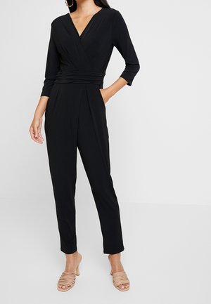 Kvinde iført en sort, omslagsstil jumpsuit med trekvartærmer og fremhævet talje, kombineret med beige åbne hælede sandaler.