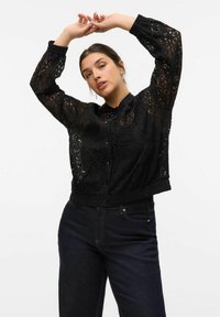 Veste courte en dentelle noire avec un devant à boutons, des manches longues bouffantes et un motif floral texturé, associée à un jean en denim foncé.