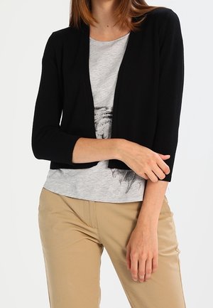 Vrouw die een zwarte open vallende cardigan draagt over een grijs t-shirt met opdruk, gecombineerd met beige broek, tegen een effen witte achtergrond.