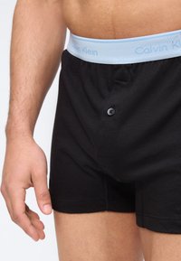 Κλείσιμο ενός ανδρικού κορμού και χεριού που φοράει μαύρα boxer εσώρουχα με μπλε ζώνη μέσης Calvin Klein και εμπρόσθιο άνοιγμα με κουμπιά.