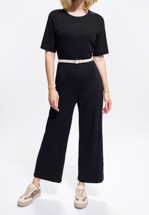 Frau trägt einen schwarzen kurzärmeligen Jumpsuit mit einem hellbeigen Gürtel und beigen Sneakers mit Leopardenmuster-Akzenten.
