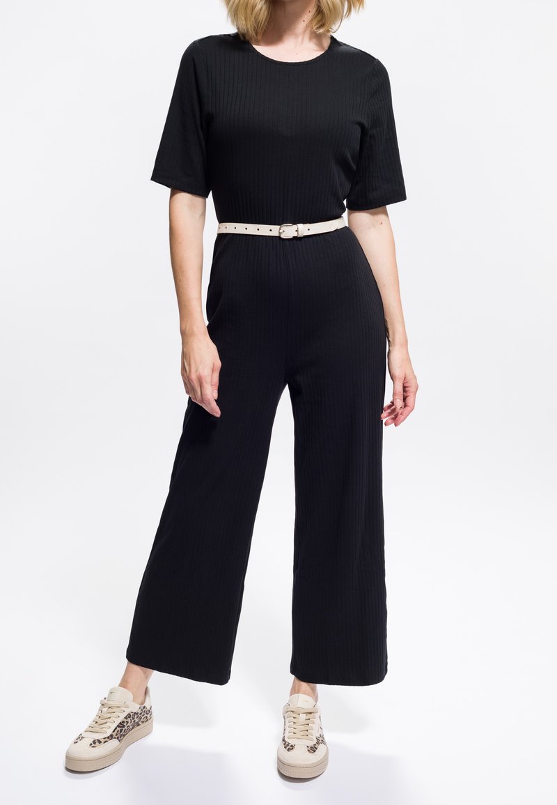 Vrouw in een zwart kortmouwen jumpsuit met een lichtbeige riem en beige sneakers met luipaardprintaccenten.
