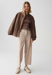 Giacca bomber in pile marrone sopra una maglia marrone, abbinata a pantaloni larghi di un beige chiaro e sneakers beige con accenti marroni.