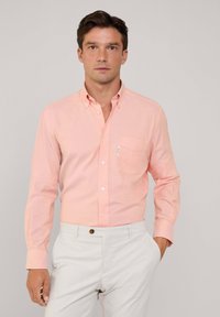 Camisa de botones de color rosa claro con mangas largas, un bolsillo en el pecho y botones blancos, combinada con pantalones de color claro. Textura sólida.