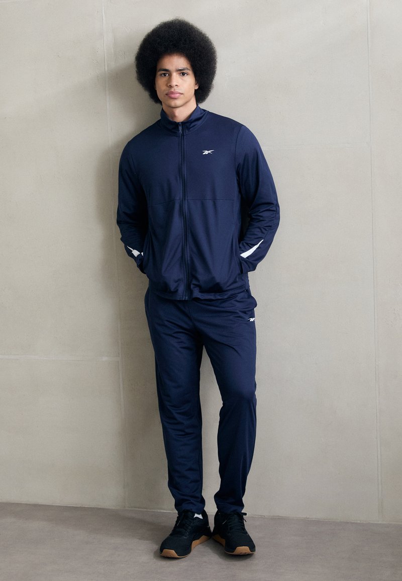 Chándal deportivo azul marino confeccionado en tejido suave, que incluye una chaqueta con cremallera y detalles en blanco, y pantalones a juego, combinados con zapatillas negras.