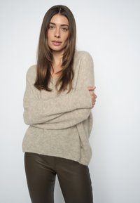 Femme aux longs cheveux bruns portant un pull en tricot beige et un pantalon marron foncé, debout les bras croisés devant un fond uni.