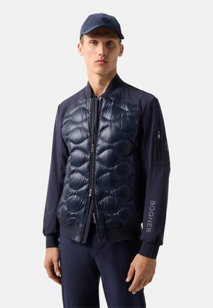 Man draagt een marineblauwe doorgestikte bomberjas met ritssluitingen en bijpassende marineblauwe pet tegen een effen achtergrond.