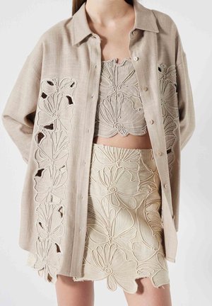 Beiges Leinenoutfit beinhaltet ein oversized Hemd, ein kurzes Top und einen Rock mit großen floralen Cut-Out-Stickmustern.