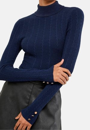 Personne portant un pull à col roulé côtelé bleu marine avec des boutons dorés sur les manches et une jupe en cuir noire, bras croisés.