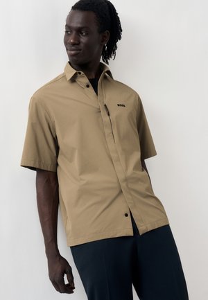 Άντρας με dreadlocks που φοράει μια κοντομάνικη μπεζ μπλούζα με κουμπιά και το λογότυπο "BOSS" καθώς και σκούρο παντελόνι, στέκεται μπροστά σε ανοιχτό φόντο.