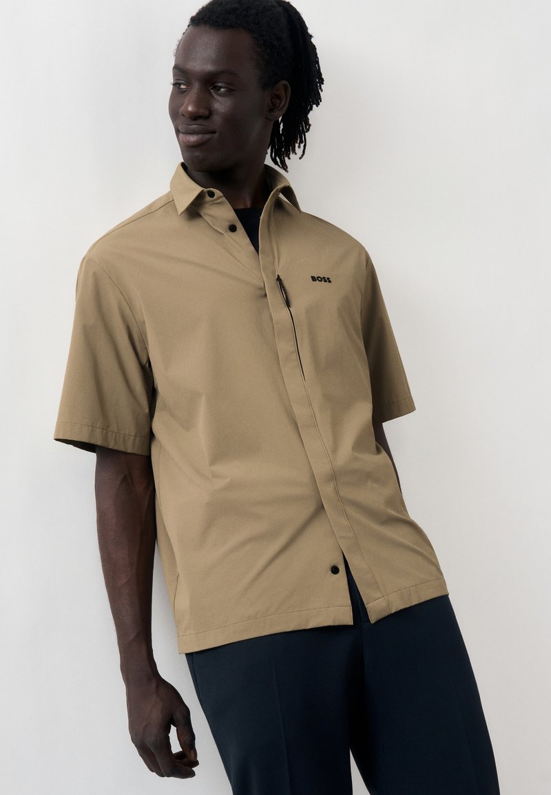 Άντρας με dreadlocks που φοράει μια κοντομάνικη μπεζ μπλούζα με κουμπιά και το λογότυπο "BOSS" καθώς και σκούρο παντελόνι, στέκεται μπροστά σε ανοιχτό φόντο.