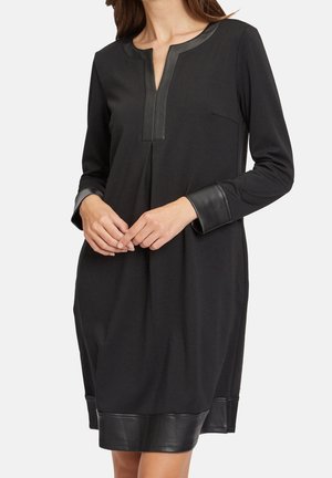 Schwarzes langärmeliges Kleid mit Lederbesatz an Kragen, Manschetten und Saum, getragen von einer Person mit gefalteten Händen vorne.