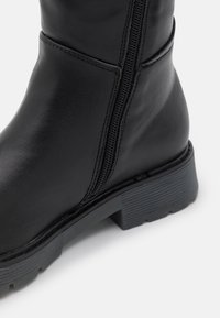 Bottine noire en cuir lisse avec une fermeture éclair sur le côté, semelle en caoutchouc robuste et un léger motif texturé sur le talon pour une meilleure adhérence.