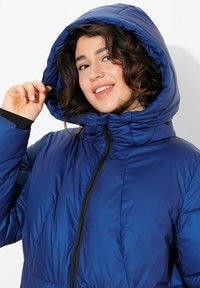 Niebieska insulated puffer jacket z zapięciem na zamek i dużym kapturem. Materiał ma błyszczące wykończenie i charakteryzuje się pikowaną teksturą.
