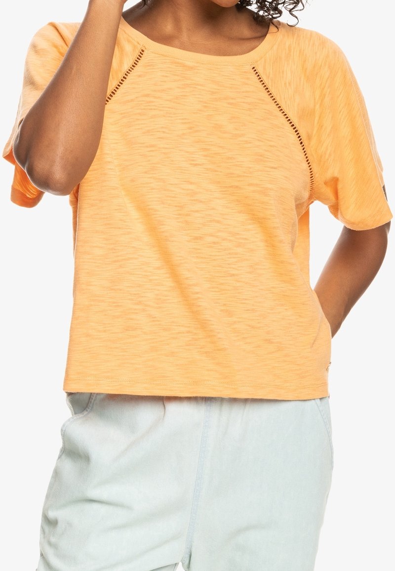 Krátký rukáv, oranžové tričko s texturovaným povrchem a raglánovými rukávy. Obsahuje malé výřezy podél švů pro ventilaci.