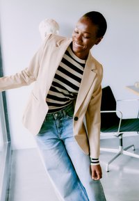 Lächelnde Frau, die seitlich am Fenster lehnt, trägt einen beigen Blazer, ein schwarz-weiß gestreiftes Hemd und hellblaue Jeans in einem hellen, modernen Raum.