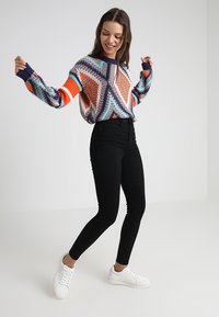 Multikleur gebreide sweater met een geometrisch patroon in oranje, blauw en marineblauw. Gecombineerd met hoge, zwarte jeans en witte sneakers.