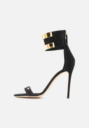 Sort ruskind stiletto sandal med tynd ankelrem med en bred guldfarvet metal manchet og et lille gyldent logo på den forreste rem.