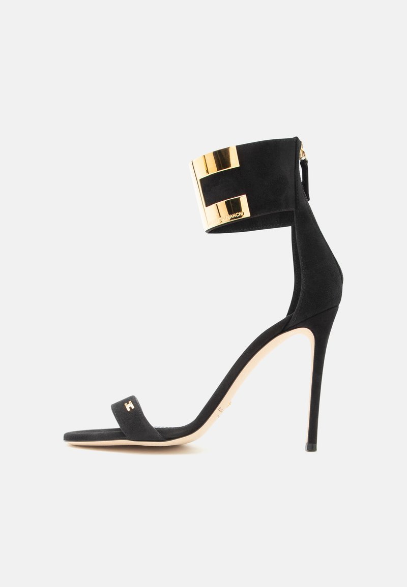 Μαύρο σουέτ πέδιλο στιλέτο με λεπτό λουράκι στον αστράγαλο που διαθέτει φαρδύ μεταλλικό χρυσό μανσέτα και μικρό χρυσό λογότυπο στο μπροστινό λουράκι.