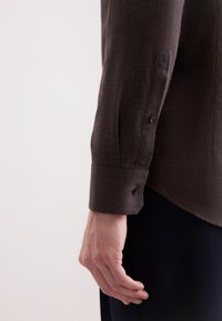 Camicia marrone a maniche lunghe con tessuto strutturato. Presenta polsini con bottoni e un orlo curvato. Nessun motivo o decorazione visibile.