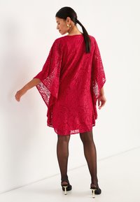 Röd spetskaftan-klänning med breda ärmar och ett mönstrat tyg. Bärs över svarta strumpbyxor och matchas med högklackade sandaler.