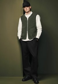 Green knit vest with two front pockets over a white button-up shirt, paired with loose black trousers and black lace-up shoes. Black cap worn.