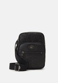 Sort crossbody taske med struktureret geometrisk mønster, lynlåslomme foran og justerbar rem. Lacoste-logoet er præsenteret på forsiden.