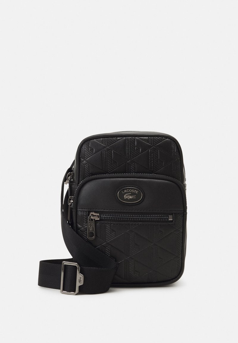 Mala de ombro preta com padrão geométrico texturizado, bolso frontal com fecho, e alça ajustável. Logotipo da Lacoste em destaque na frente.