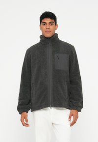 Vīrietis valkā tumši pelēku flīsa jaku ar priekšējo rāvējslēdzēju un krūšu kabatu, apvienojot to ar baltām biksēm, stāvot pret vienkāršu fonu.