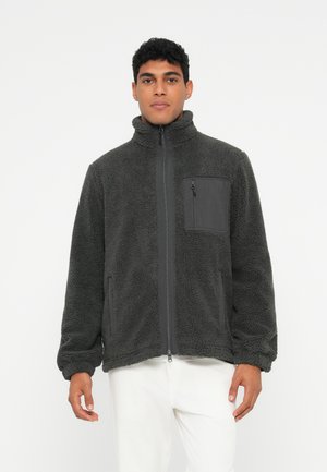 Man med en mörkgrå fleecejacka med framdragkedja och bröstficka, i kombination med vita byxor, står mot en enkel bakgrund.