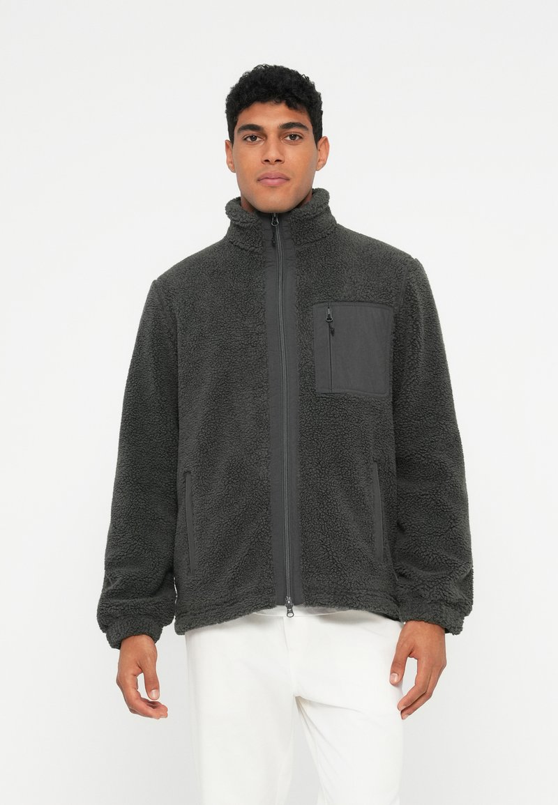 Vīrietis valkā tumši pelēku flīsa jaku ar priekšējo rāvējslēdzēju un krūšu kabatu, apvienojot to ar baltām biksēm, stāvot pret vienkāršu fonu.