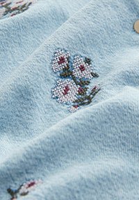 Teren de denim albastru deschis cu flori albe brodate, având accente roșii. Textură netedă cu un model de cusătură vizibil și un detaliu cu nasture.