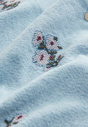 Tissu en denim bleu clair avec des fleurs blanches brodées comportant des accents rouges. Texture lisse avec un motif de couture visible et un détail de bouton.