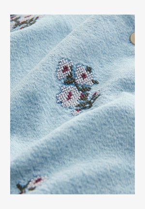 Svetlomodrá denimová látka s vyšitými bielymi kvetmi, ktoré majú červené akcenty. Hladká textúra s viditeľným šitím a detailom v podobe gombíka.