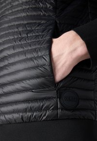 Μαύρο επενδυμένο μπουφάν με υφή και σχεδίαση καπιτονέ. Διαθέτει θηλάκι με φερμουάρ και κυκλικό λογότυπο στην αριστερή πλευρά.