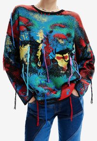 Pull oversized coloré avec un motif vibrant en rouge, bleu et vert. Présente de longs pompons aux manches et un col noir côtelé.