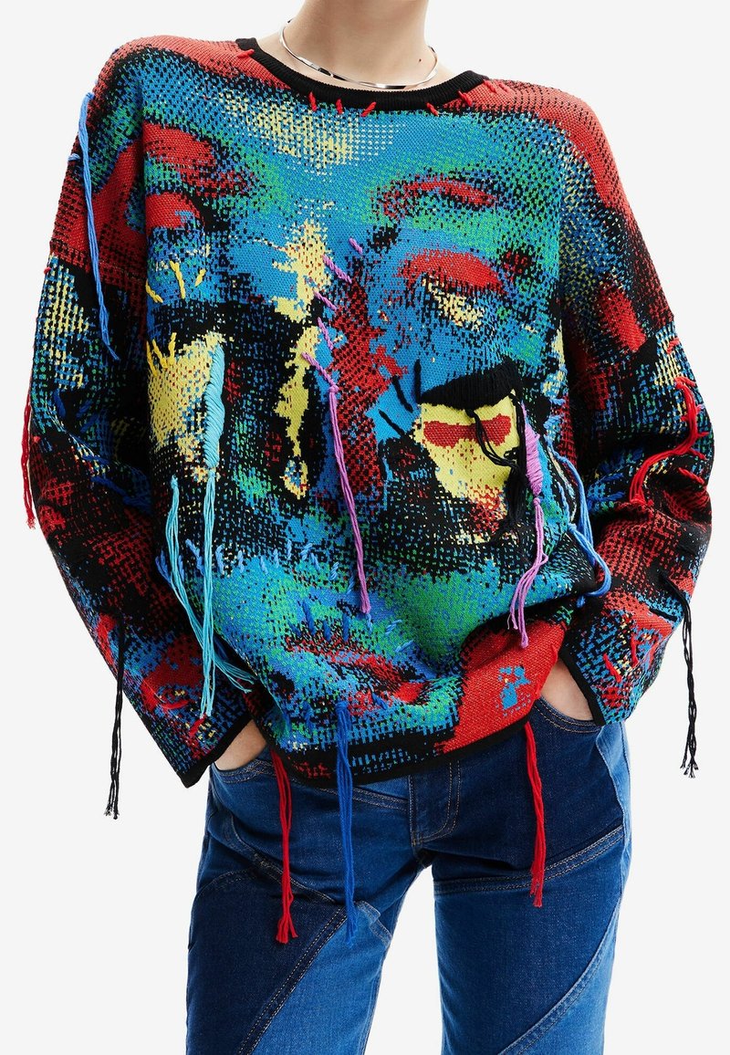 Pull oversized coloré avec un motif vibrant en rouge, bleu et vert. Présente de longs pompons aux manches et un col noir côtelé.