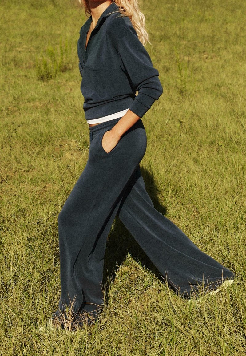Personne marchant sur l'herbe portant un pantalon bleu foncé ample et un haut assorti à manches longues avec une main dans la poche.