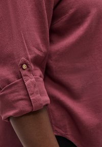 Pööratud pikad varrukad tumepunasel särgil, kinnitatud väikese pruuniga nupuga tumedale nahale.