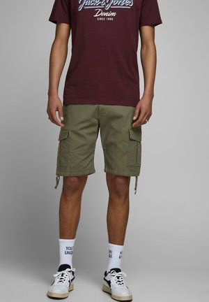 Mann trägt ein kastanienbraunes Jack & Jones T-Shirt, olivgrüne Cargo-Shorts, weiße Socken mit Schriftzug und weiße Sneaker vor grauem Hintergrund.