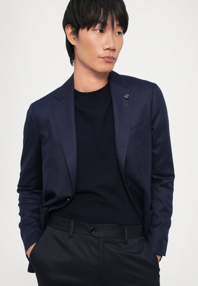 Navy blazer made of smooth fabric with a slim fit, featuring a decorative lapel pin, worn over a dark sweater and matching pants.