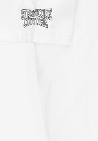 Biały bawełniany t-shirt z napisem "STREETCORE COUTURE" wytłoczonym grubymi czarnymi literami na lewej stronie klatki piersiowej; miękka faktura, krótkie rękawy.