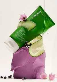 Tubo verde con un tappo bianco, etichettato "Maschera per pori ghiacciata al viola". Appoggiato su argilla viola, decorato con fette di cetriolo e fiori rosa.