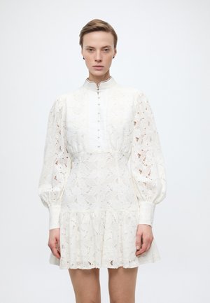 Γυναίκα που φοράει λευκό δαντελένιο φόρεμα με μακριά φουσκωτά μανίκια, ψηλό γιακά, κουμπωτό μπροστινό μέρος και βολάν στο τελείωμα, στέκεται μπροστά σε έναν απλό φόντο.